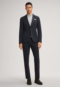 Marineblauer, maßgeschneiderter Anzug aus strukturiertem Stoff mit Reverskragen, zwei Fronttaschen und passenden Hosen. Kombiniert mit einem grauen Oberteil.