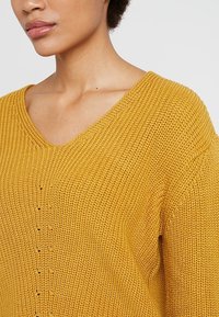 Musztardowożółty sweter z dzianiny o dekolcie w serek, luźnym kroju i fakturą z pionowymi przeszyciami na przodzie.