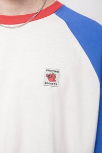 Bílá košile s krátkým rukávem a modrými raglánovými rukávy, červeným límcem a nášivkou "KAOTIKO SOCIETY" s kreslenou postavičkou. Materiál: bavlna.