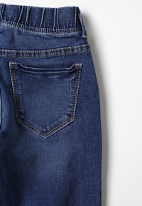 Detailní záběr na tmavomodré denimové kalhoty s elastickým pasem a prošívanou zadní kapsou na bílém pozadí.