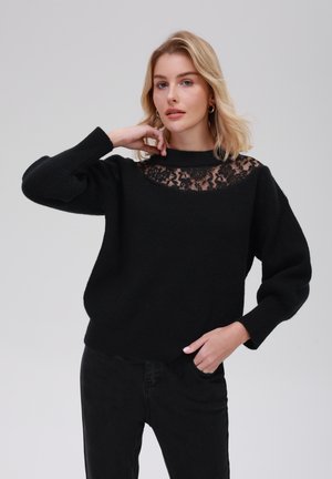 Pull en tricot noir avec un col haut, présentant un panneau en dentelle au niveau de la poitrine. Des manches surdimensionnées et une coupe décontractée achèvent le design.