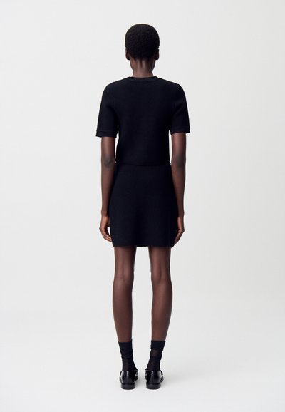 Femme de dos, portant un haut noir à manches courtes et une jupe courte assortie, avec des chaussettes et des chaussures noires, sur un fond uni.