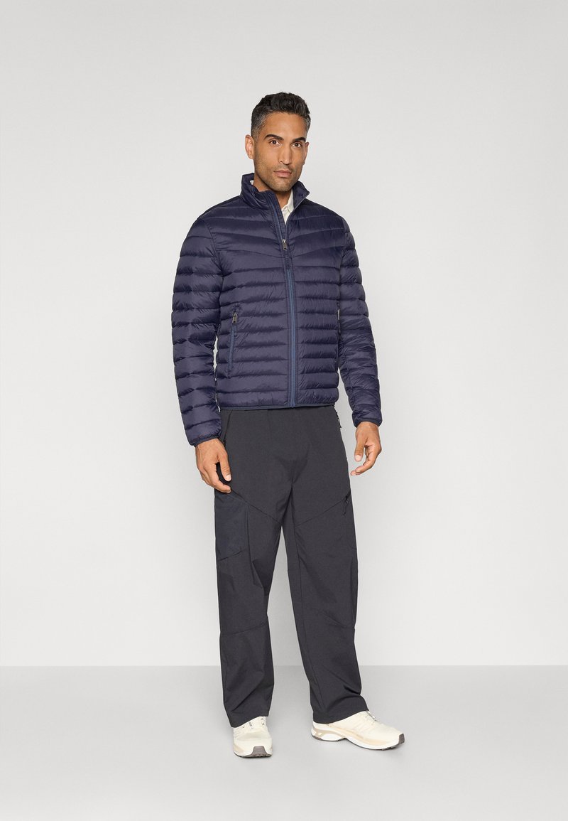 Marineblauer Daunenjacke mit horizontaler Steppung, hohem Kragen und Reißverschluss, kombiniert mit dunklen Hosen und weißen Sportschuhen.