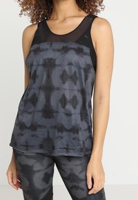 Kvinna som bär en ärmlös atletisk topp i svart och grått tie-dye med mesh-krage och matchande leggings, stående mot en enfärgad bakgrund.