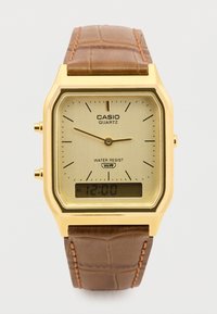 Złoty kwadratowy zegarek Casio na kwarc, z beżową tarczą, czarnymi oznaczeniami i wyświetlaczem cyfrowym. Brązowy skórzany pasek w wzór krokodyla.