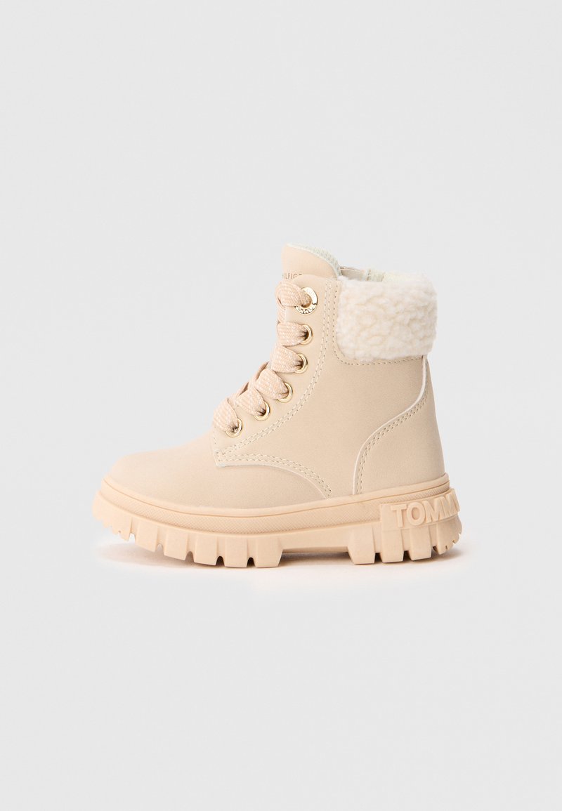 Beige ankelstøvel med tykk gummisåle, polstret ankelkrage foret med hvit fleece, og beige snørebånd med gulløyestykker.
