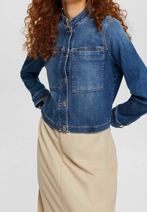 Femme aux cheveux roux bouclés portant une veste en denim bleue boutonnée et une jupe beige, les mains reposant sur les hanches devant un fond uni.