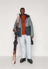 Smučarska obleka sivo-tirensko jakno, oranžno majico, belimi hlačami in črnimi smučarskimi čevlji. Moški drži črne smuči z rdečimi poudarki.