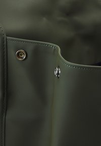 Matériau imperméable vert présentant deux œillets métalliques et des coutures renforcées le long des bords, avec une texture lisse et mate.