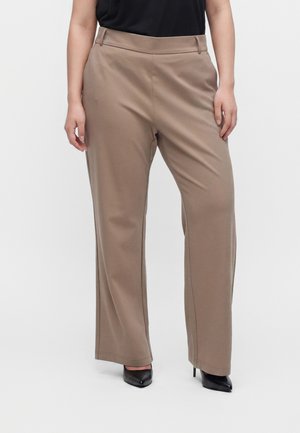 Femme portant un pantalon large couleur taupe et des talons hauts noirs pointus, debout devant un fond uni clair.