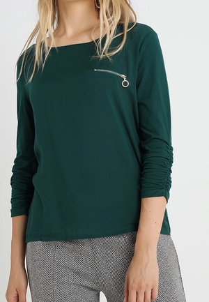 Femme portant un haut à manches longues vert foncé avec un petit détail de fermeture éclair horizontale sur la poitrine et un pantalon gris à motif chevron.