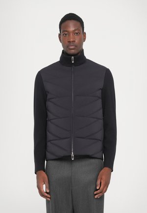 Black padded vest with a high collar, front zipper, and chevron quilted pattern, layered over a black long-sleeve shirt and gray trousers.
