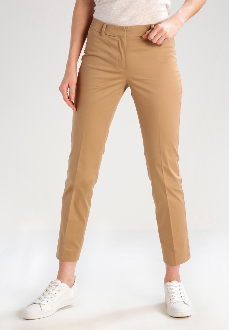Femme portant un pantalon camel ajusté et des baskets blanches, debout devant un fond clair uni.