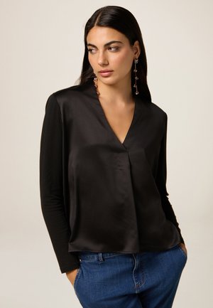 Maglietta nera a maniche lunghe con scollatura a V, realizzata in morbido tessuto satinato, con una vestibilità comoda e tasche laterali, abbinata a jeans blu.