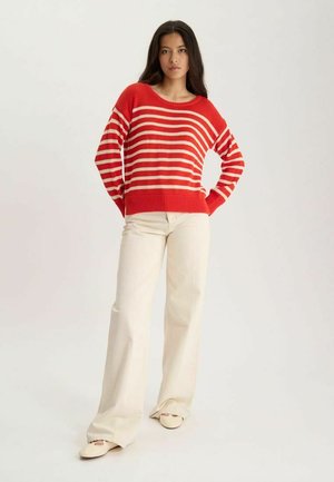 Femme debout, les mains sur les hanches, portant un pull rayé rouge et beige, un pantalon crème à jambes larges et des chaussures beiges sur un fond uni.