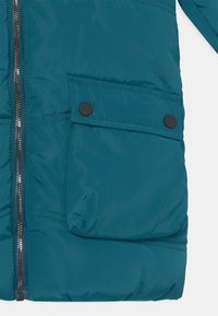 Türkise gepolsterte Jacke mit großem Klappentasche, die mit zwei schwarzen Druckknöpfen gesichert ist, und sichtbarem silbernen Reißverschluss auf der linken Seite.
