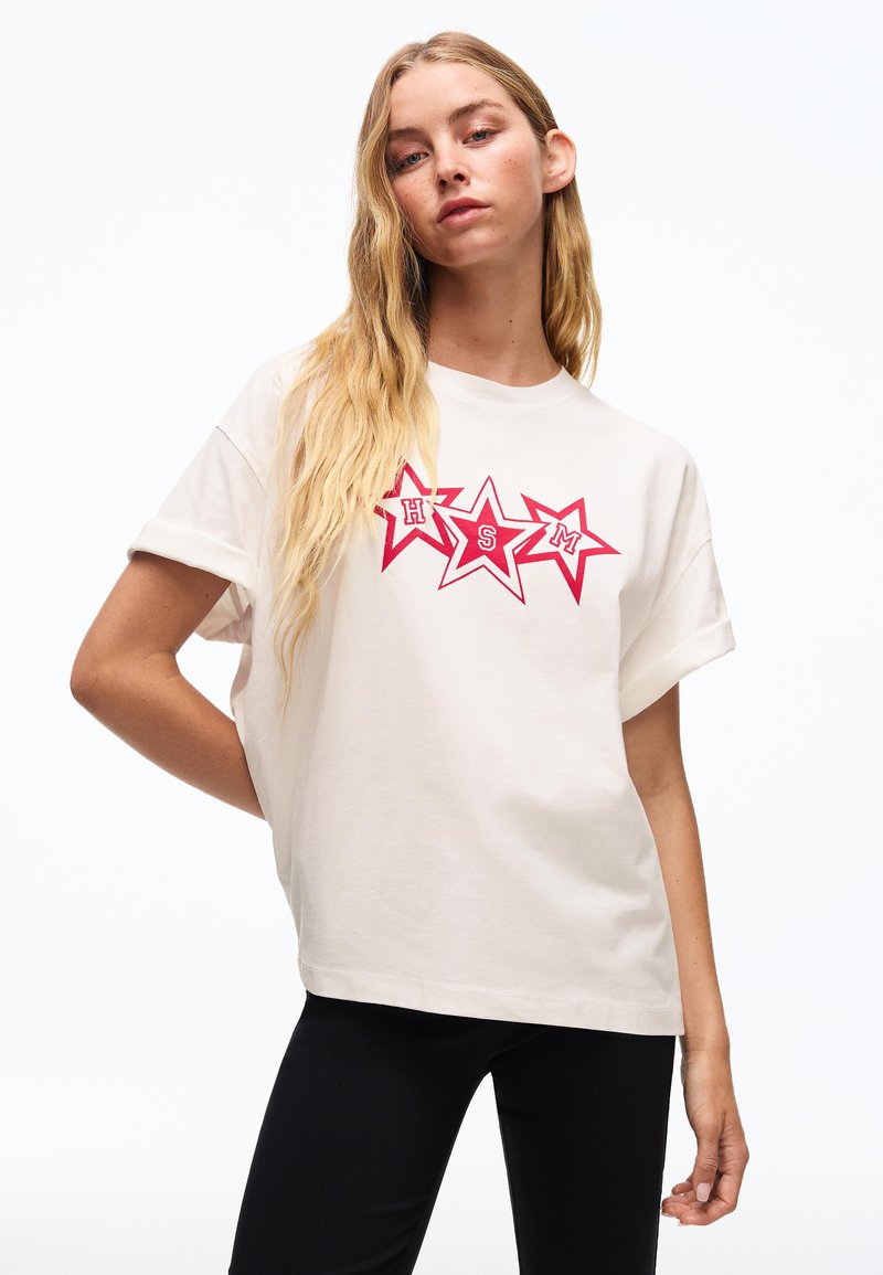 Jeune femme aux longs cheveux blonds portant un T-shirt blanc avec trois étoiles rouges portant les lettres H, S, M, associé à un pantalon noir.