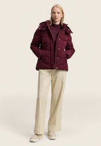 Grimizna jakna na preklop s kapuljačom, prednjim zatvaračem na patentni zatvarač i dva džepa. Uparena s kremasto bijelim hlačama i svijetlim čizmama.