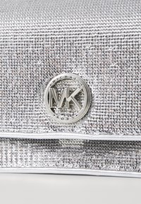 Srebrna kopertówka zdobiona koralikami o gładkiej fakturze. Posiada okrągłe logo MK w metalicznym srebrnym kolorze. Płaski design z zamkiem błyskawicznym.