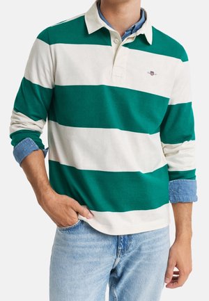 Mann trägt ein grünes und weißes Rugbyshirt mit breiten Streifen über einem blauen Chambray-Hemden mit Knöpfen und hellblauen Jeans, eine Hand in der Tasche.