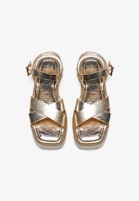 Gullmetalliske sandaler med kryssede stropper, flat såle og ankelspenne. Glatt tekstur med en blank finish og firkantet tådesign.