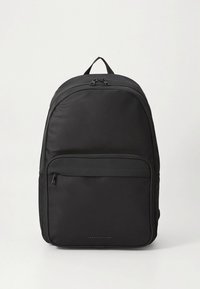 Svart nylonsekk med en glatt tekstur, avrundet topp, frontlomme med glidelås og en logo nederst. Justerbare polstrede stropper.