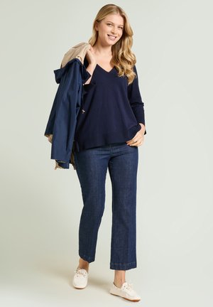 Smiling woman in navy V-neck sweater and dark jeans holds a beige and navy jacket over her shoulder, wearing white sneakers.