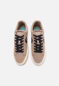 Beżowe sneakersy z płótna, z czarnymi sznurowadłami, niebieskim okrągłym wzorem na wkładce i czarnym logo w kształcie gwiazdy po bokach. Białe gumowe podeszwy.