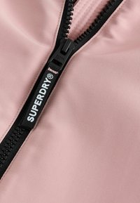 Chaqueta de color rosa claro con cremallera y etiqueta con el logo 'SUPERDRY'. Tejido texturizado con superficie lisa y detalle de cremallera.