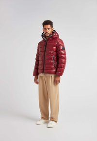 Rød pufferjakke med en skinnende finish, lynlås foran og hætte. Har sort logodetalje og quiltet mønster. Kombineret med beige bukser og hvide sneakers.