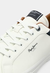 Sneaker alb de la Pepe Jeans cu sireturi albe, logo negru pe lateral și accente maro în jurul gulerului și etichetei de pe limbă.