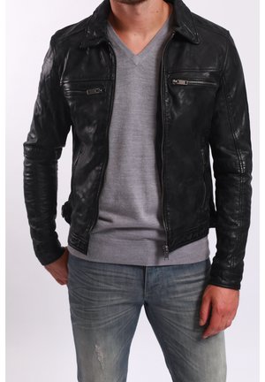 Mężczyzna ubrany w czarną skórzaną kurtkę na szarym swetrze z dekoltem w serek i wyblakłych niebieskich dżinsach, stojący z rękami wzdłuż ciała.