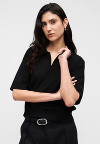 Μαύρο πόλο με κοντά μανίκια και φερμουάρ στον γιακά, μαλακό ύφασμα και εφαρμοστή κάτω ραφή. Συνδυάζεται με μαύρο παντελόνι και ζώνη με ασημένια αγκράφα.