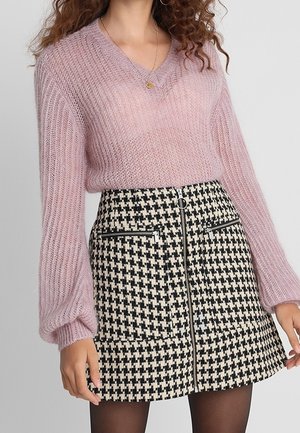 Femme portant un pull en maille rose pelucheuse et une jupe à motif pied-de-poule noir et blanc avec des détails zippés.