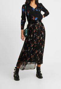 Zwarte blouse met lange mouwen en bloemenprints, gecombineerd met een geplooide zwarte rok met rode en witte bloemen. Zwarte combat boots en een groene tas.