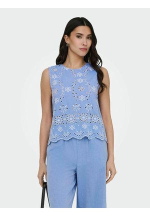 Femme portant un haut sans manches bleu clair avec une broderie florale blanche et un pantalon assorti, tenant un sac à main noir à ses côtés.