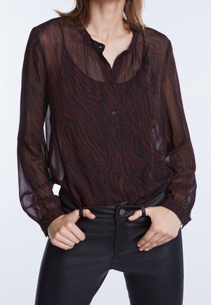 Femme portant un chemisier transparent à manches longues, à motif sombre, rentré dans un pantalon en cuir noir taille haute, les mains dans les passants de ceinture.