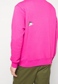 Svetle růžová mikina s kulatým výstřihem a žebrovanými manžetami. Má malé logo Nike v černé a bílé barvě na zádech poblíž ramene.