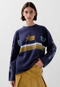 Sudadera de manga larga en color marino con diseño de estrella dorada y el número 18. Presenta rayas horizontales en blanco y dorado, acentos en azul y detalles gráficos.
