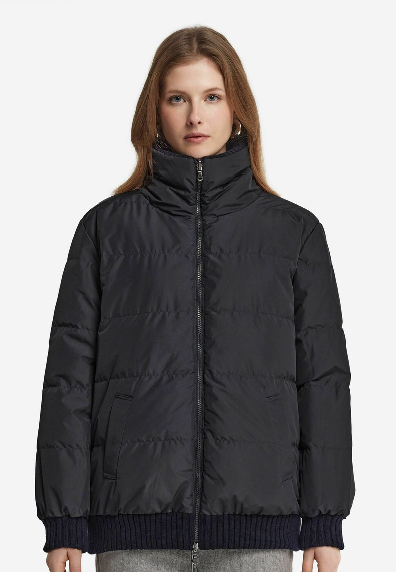 Svart puffjacka med hög krage, dragkedja fram och sidofickor. Har ett kviltat mönster och ribbade marinblå muddarna vid ärmsluten.