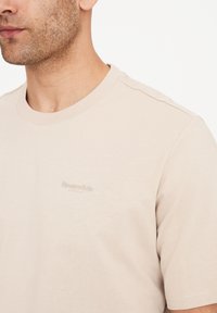 Lysbeige kortærmet t-shirt lavet af blødt stof, med et subtilt præget logo på brystet og en klassisk rund halsudskæring.
