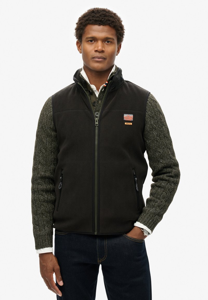 Gilet en polaire noir avec des manches texturées vertes, fermeture zippée à l'avant et poches. Il possède un patch logo sur la poitrine gauche. Design épuré et décontracté.