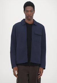 Navy blue jacket with a button-up collar, single chest pocket, and a textured fabric. Worn over a black shirt and brown pants.