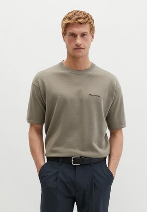 Blondyn noszący taupe krótkodruki sweter z logo "Marc O'Polo" i granatowe spodnie, ręce w kieszeniach, neutralne tło.