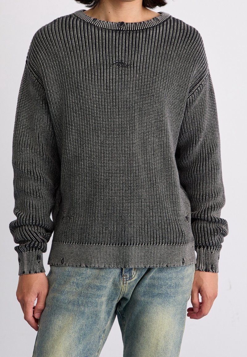 Personne portant un pull en maille texturée noir et blanc avec poignets et ourlet côtelés, associé à un jean bleu délavé.