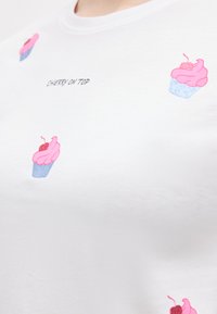 Λευκό T-shirt με ροζ εικονογραφήσεις cupcakes, διακοσμημένα με κόκκινες κερασιές, και το κείμενο «CHERRY ON TOP» σε μαύρη καλλιγραφική γραμματοσειρά στο στήθος.