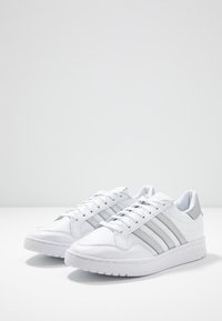 Vita atletiska skor med en slät läderöverdel, som har tre silverränder, en vadderad krage och en texturerad gummisula.