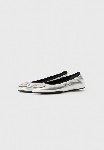 Une paire de ballerines métalliques argentées avec des bords noirs et un emblème rond décoratif argenté sur chaque orteil, présentée sur un fond blanc.