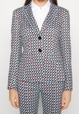 Vrouw draagt een nauwsluitend blazer en broek met een blauw, rood en zwart driehoekig geometrisch patroon over een wit overhemd met kraag.