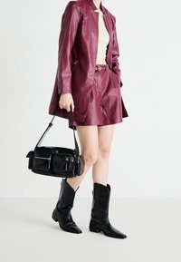 Burgundy konstläderblazer, matchande shorts, svart handväska med dragkedjor, och svarta spetsiga cowboystövlar mot en ljus bakgrund.